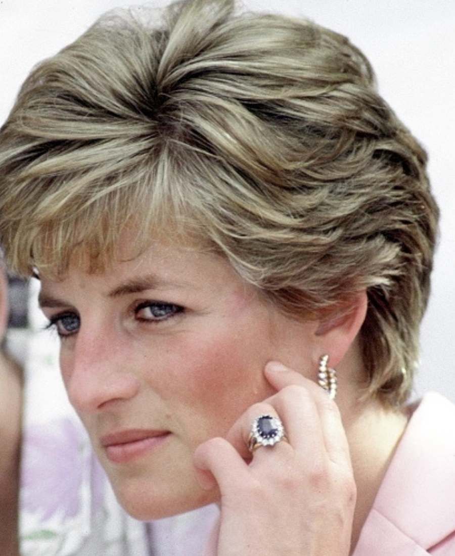 Foto de rosto da princesa de Gales em um lugar público. Ela está com a mão no rosto e usa anel de noivado e brinco.