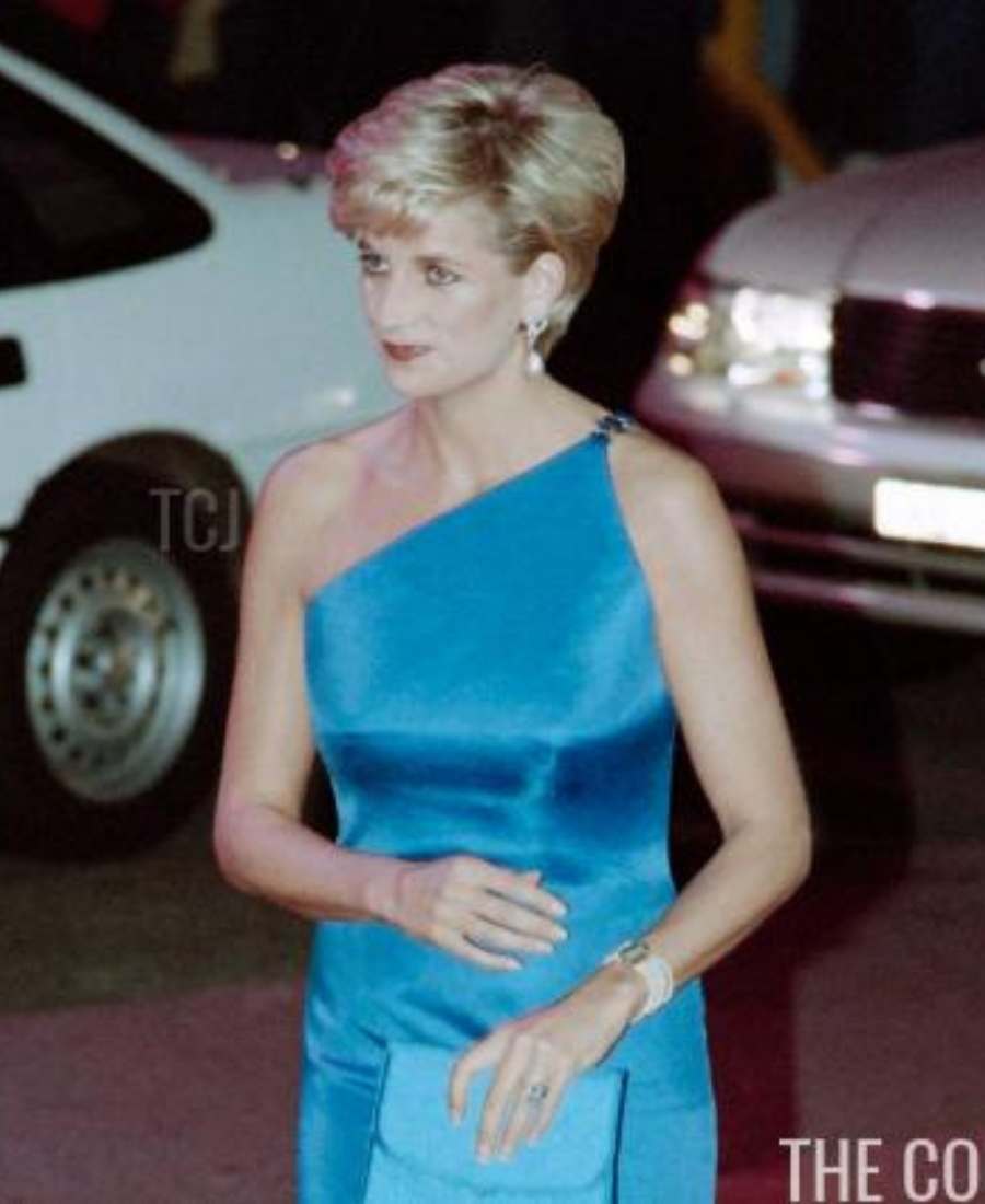 Foto de lady Di em local público com dois carros. Ela está de frente, em pé e usa anel e pulseira água-marinha. Lady Di usa vestido azul e segura bolsa de mesma tonalidade em uma das mãos.