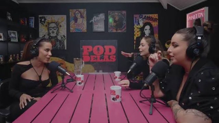 Anitta conversando com Anitta dá entrevista à Tatá Estaniecki e Bruna Unzueta em uma mesa. A cantora está de um lado e as apresentadoras do outro.