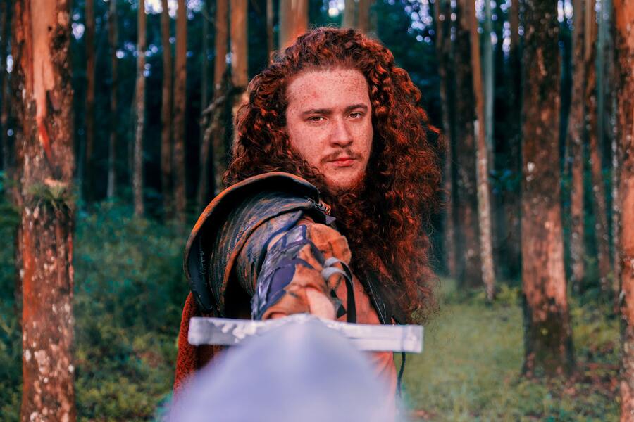 Homem do arquétipo guerreiro, ruivo, com cabelos longos e cacheados impondo uma espada em uma floresta