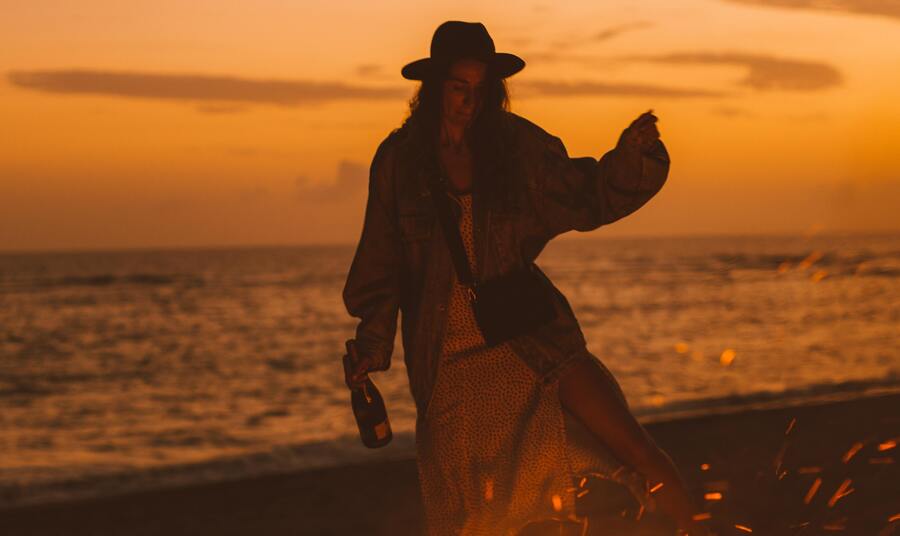 Mulher com chapéu, casaco e saia longa dançando com uma garrafa na mão na frente de uma fogueira acessa na praia