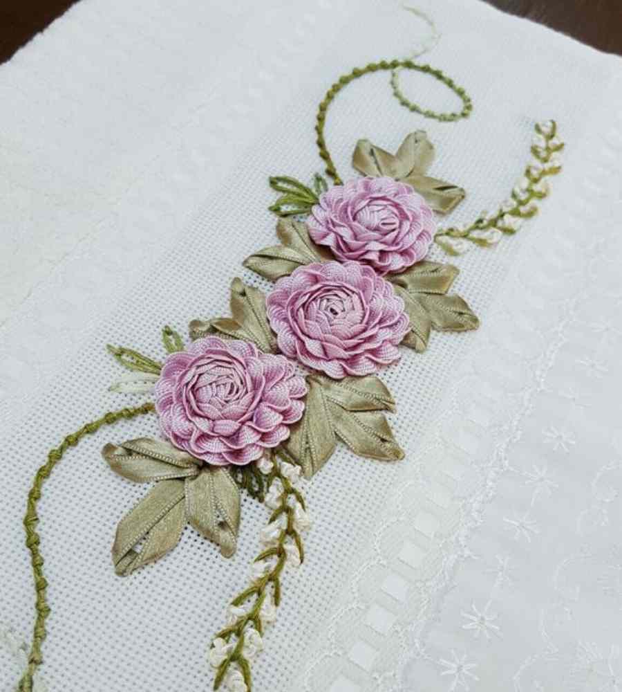 Foto toalha branca com acabamento artesanal bordado de flores roxas e folhas verdes em cetim. Peça está sobre superfície.