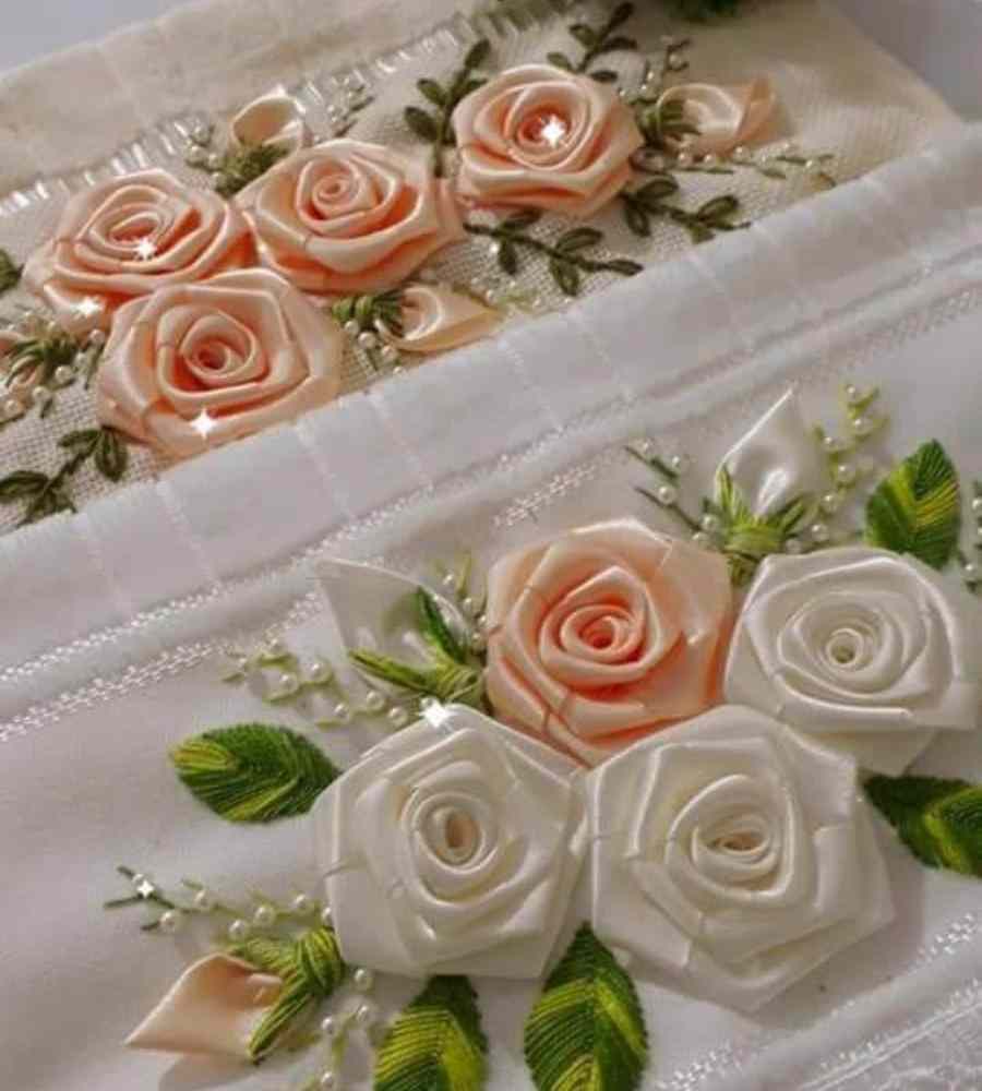 Foto toalha branca com acabamento artesanal bordado de rosas e botões brancos e bejes em cetim. Peça está sobre superfície.