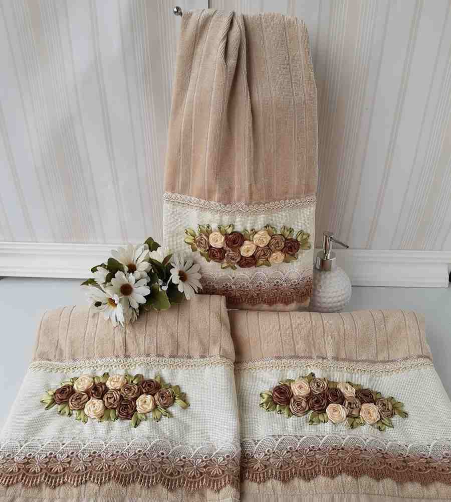 Foto toalha marrons com acabamento artesanal bordado de rosas marrons e creme em cetim. Peça está sobre superfície e penduradas em gancho ao lado de flores artificiais e porta-sabonete líquido.