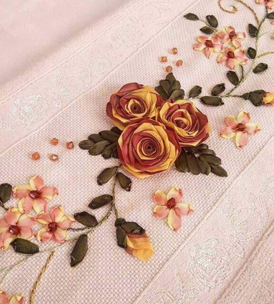 Foto toalha bege com acabamento artesanal bordado de rosas e flores em tons outonais em cetim. Peça está sobre superfície.