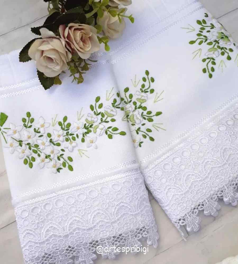 Foto toalha branca com acabamento artesanal bordado ramo de flores brancas. Peças estão sobre superfície e ao lado de flores artificiais.