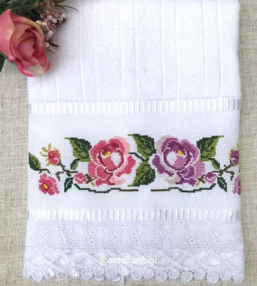 Foto toalha branca com acabamento artesanal bordado flores rosas e roxas. Peças estão sobre superfície e ao lado de flores artificiais.
