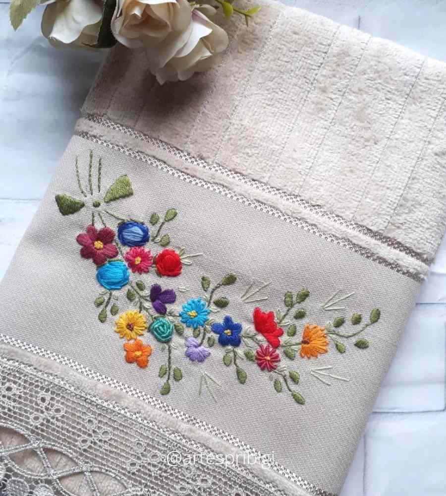 Foto toalha bege com acabamento artesanal bordado ramo de flores coloridas. Peça está sobre superfície e ao lado de flores artificiais.