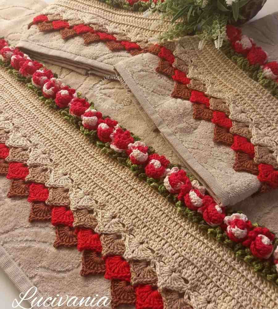 Foto peças na cor bege com acabamento artesanal em crochê marrom, vermelho e verde. As peças estão sobre superfície ao lado de rosas planta artificial verde.