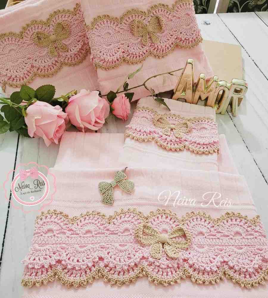 Foto de jogo completo de toalhas rosas com acabamento artesanal em crochê rosa e dourado. Peças estão sob madeira ao lado de rosas cor de rosa e item de decoração escrito a palavra amor.