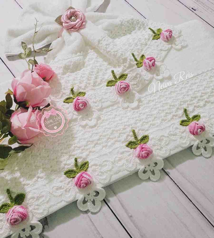 Foto jogo de toalhas brancas com acabamento artesanal em crochê branco, rosa e verde. As peças estão sobre mesa de madeira e ao lado de rosas cor de rosa.