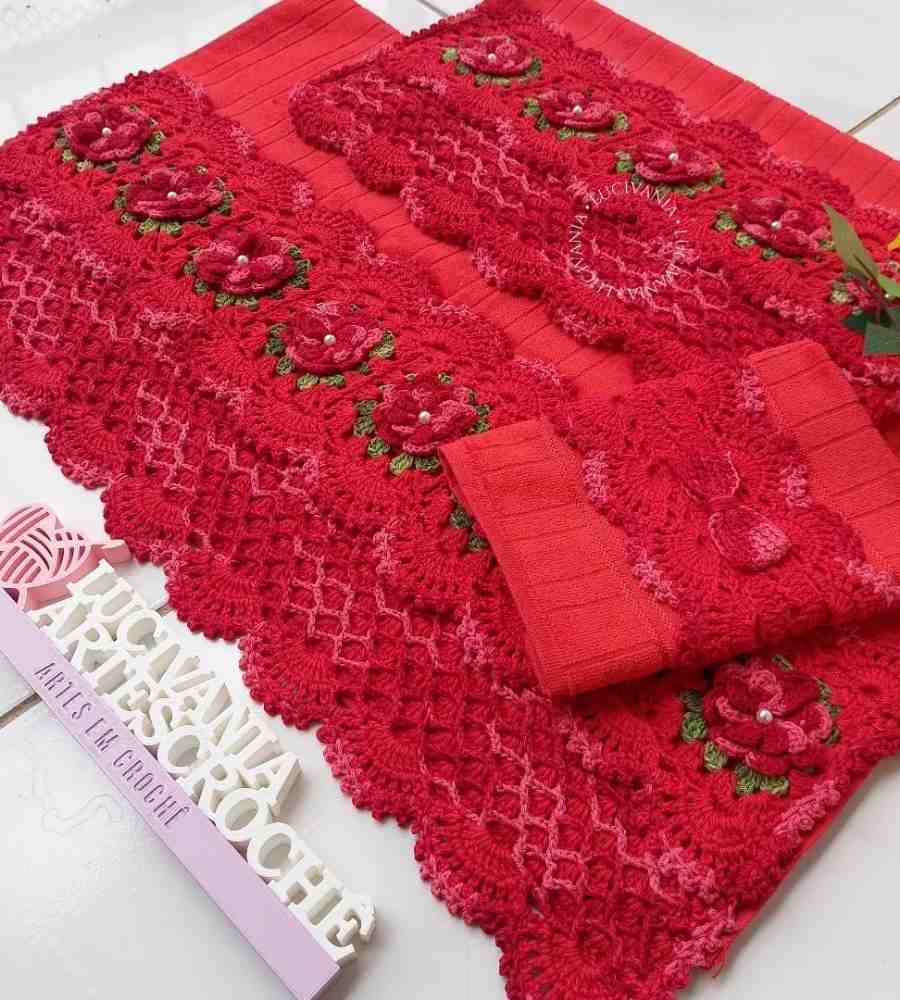 Foto jogo de toalhas vermelhas com acabamento artesanal em crochê vermelho e verde. As peças estão sobre mesa de madeira e ao lado de plaquinha com nome da artesã Lucivania Artes Crochê.