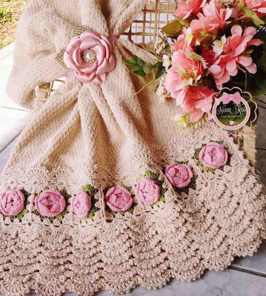 Foto de toalha de banho bege com acabamento artesanal em crochê bege, rosa e verde. A peça está no chão ao lado de flores artificiais.