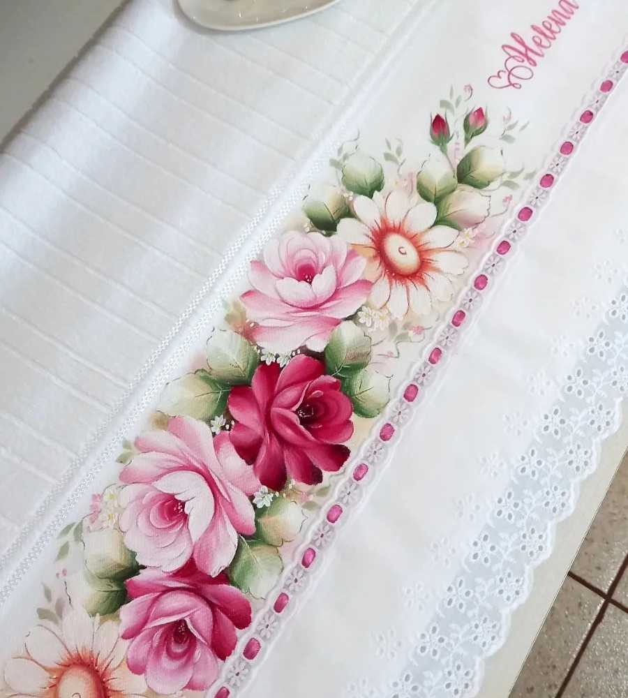 Foto toalha branca com acabamento artesanal pintura, desenho de flores e nome "Helena". Peça está sobre superfície.
