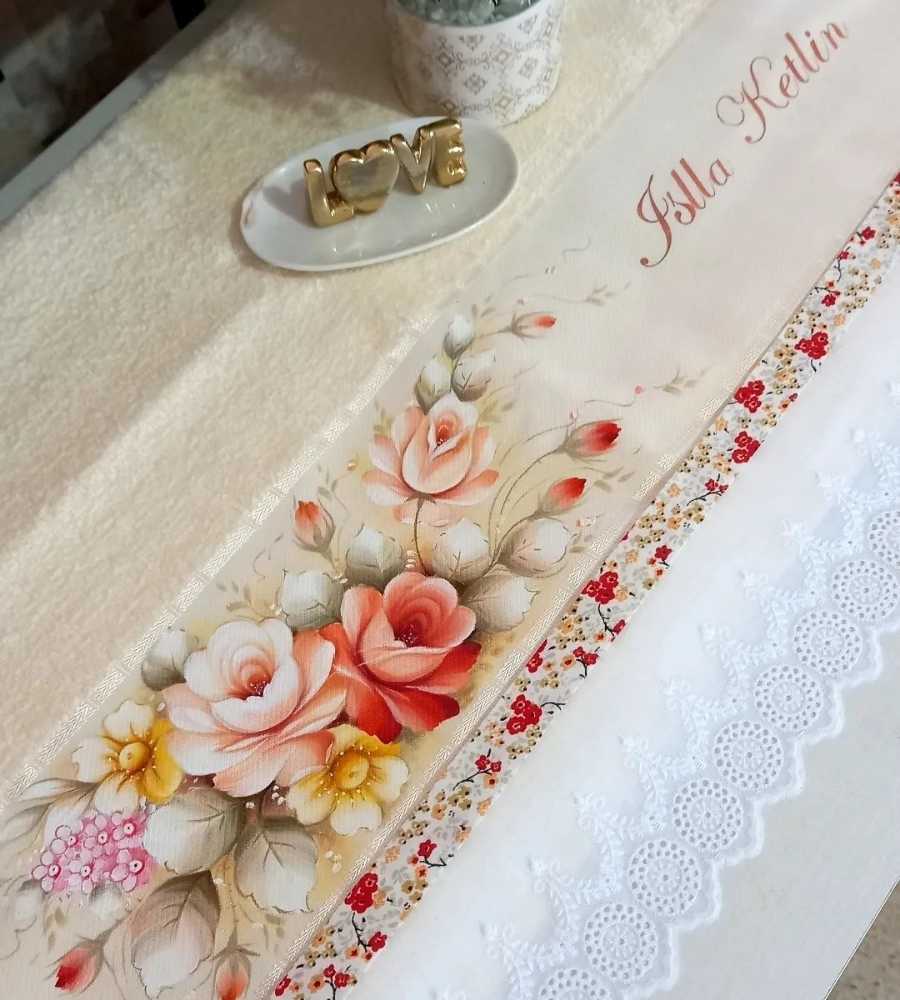 Foto toalha cor creme com acabamento artesanal pintura, desenho de flores e nome "Isla Ketlin". Peça está sobre superfície e ao lado de itens de decoração escrito "love" e platinha.