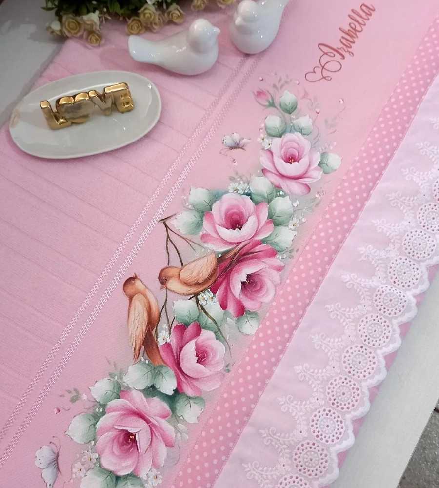 Foto toalha rosa com acabamento artesanal pintura, desenho de pássaros, flores e nome "Isabella". Peça está sobre superfície e ao lado de itens de decoração escrito "love", pássaros e flores artificias.