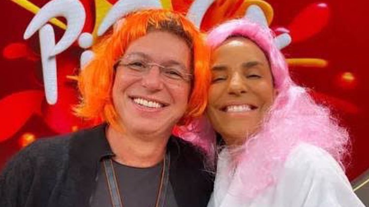 Boninho de peruca laranja e Ivete Sangalo de peruca rosa sorriem olhando para a câmera no cenário do Pipoca da Ivete.