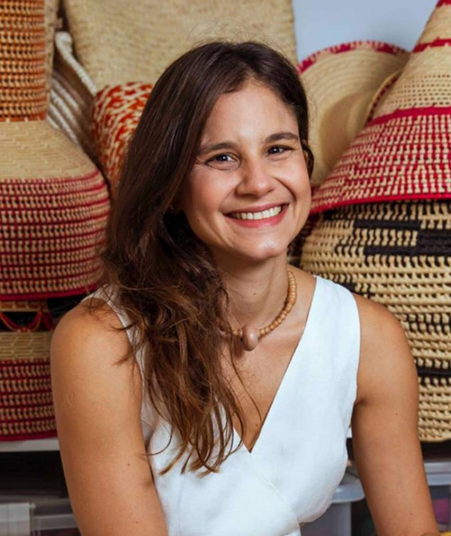 Foto de Celina Hissa, do MaxiModa 2022, sorridente. Ela usa roupa branca e esta sentada na frente de vários itens artesanais, como chapéu, feitos aparentemente de linha bege, vermelha e preta.