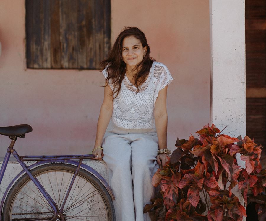 Foto de Celina Hissa sentada em muro baixo da área externa de uma casa antiga que tem janela e porta de madeira. Ela está sorridente, ao seu lado tem uma flor com folhas mescladas de marrom e vermelho e do outro lado tem a roda traseira e acento de uma bicicleta antiga de cor roxa aparecendo.