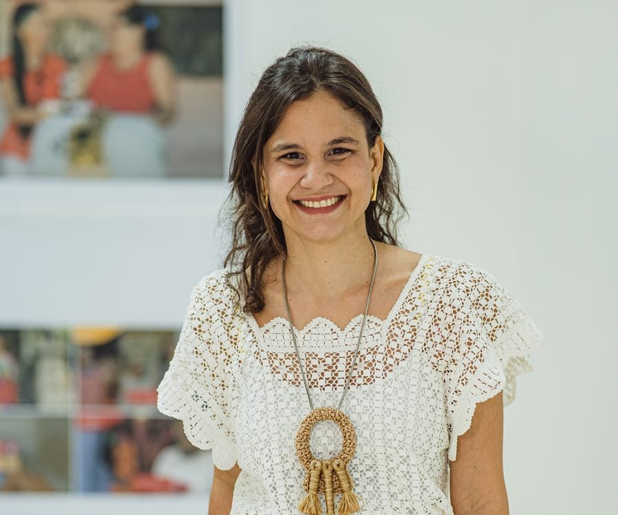 Foto de Celina Hissa, ela está sorridente, usando blusa de renda e colar artesanal. Ela está numa sala com parede branca com quadros coloridos de pessoas no fundo.
