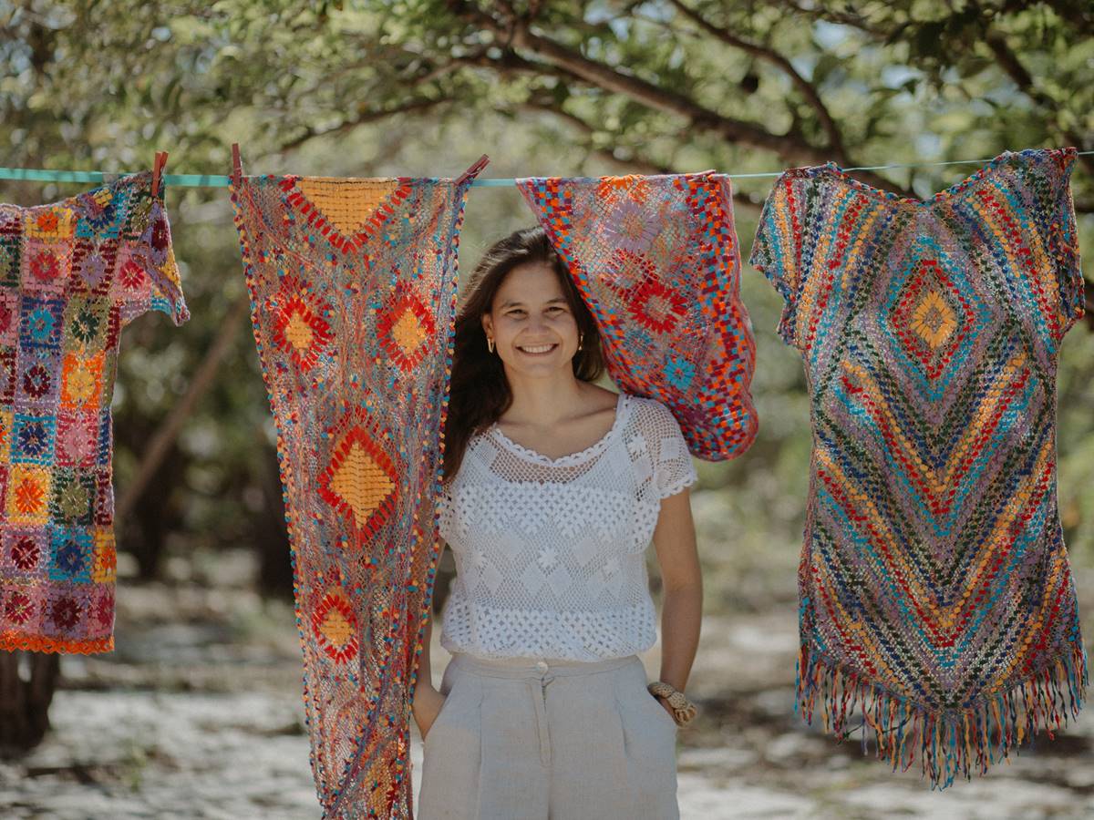 Foto de Celina Hissa de pé, sorridente, em espaço com árvores e varal com peças de roupas artesanais bastante coloridas.