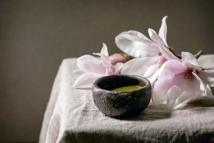 Chá de magnólia em pote marrom ao lado das flores de Magnólia em cima de mesa com toalha branca.
