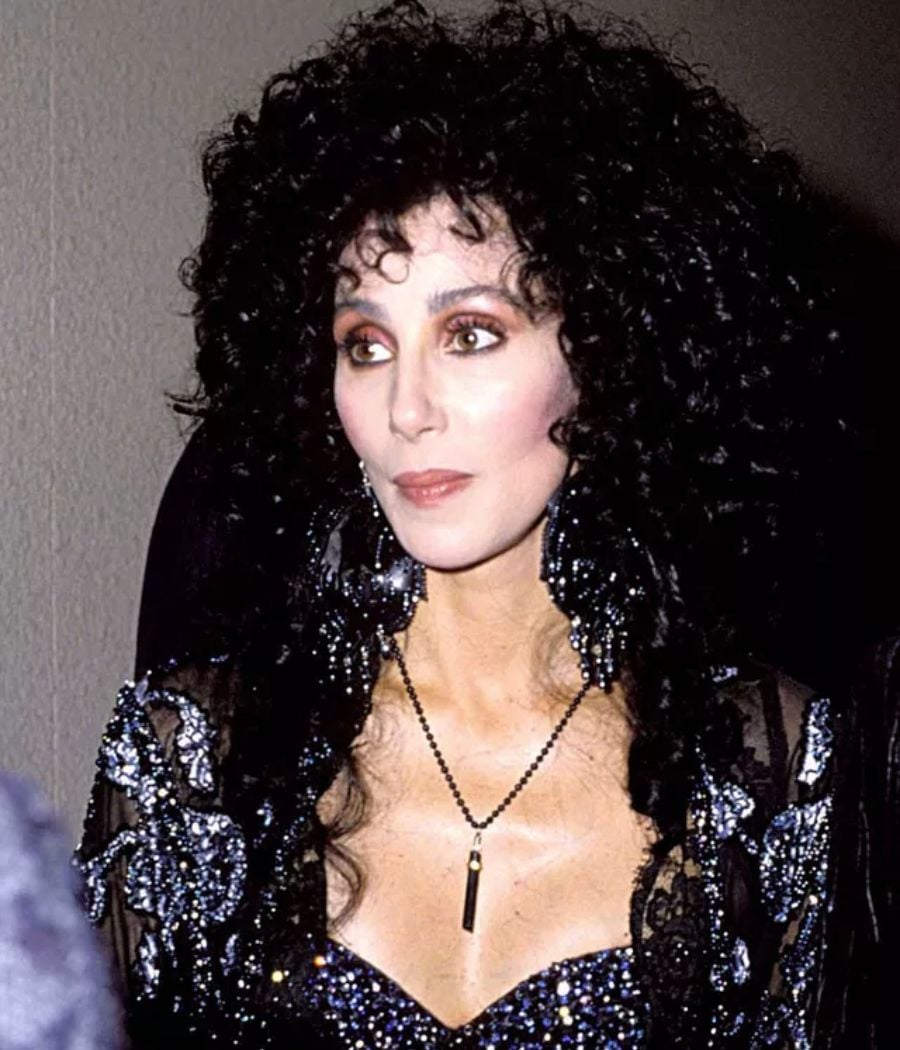 A cantora Cher com vestido preto, colar prateado e o cabelo encaracolado