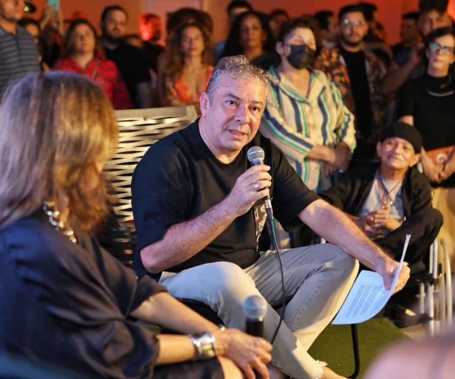 Claudio Silveira segurando microfone e papéis, sentado em cadeira ao lado de Patrícia Carta, ao redor de pessoas convidadas do coquetel de lançamento da revista Bazaar sobre o Ceará.