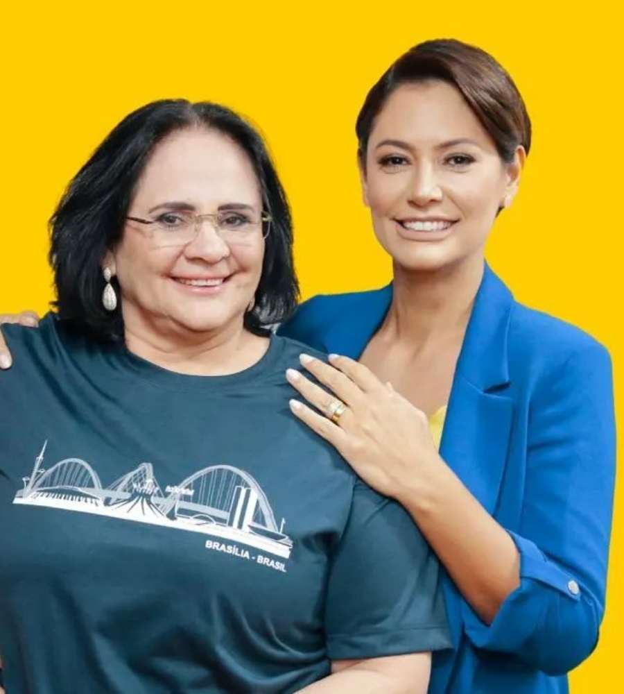 Foto de Damares Alves e Michelle Bolsonaro. Elas estão do lado uma da outra e Michelle atrás de Damares. A foto tem fundo amarelo.