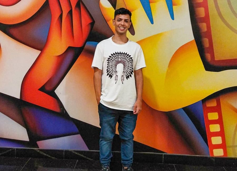 O jovem Matheus Guimarães usa uma camiseta branca e calça jeans. Ele está pousando em frente a um mural colorido