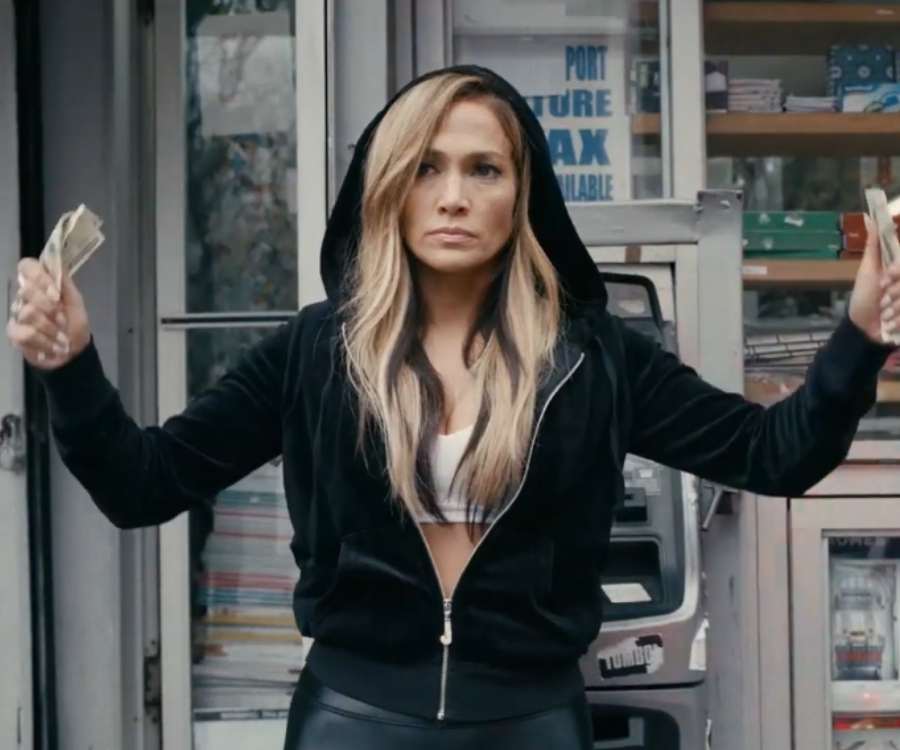 Jennifer Lopez em cena no filme "As Golpistas" em 2019. Atriz foi fotografada da cabeça ao quadril. Está com as mãos para cima segurando notas de dólares. No fundo, o que parece ser uma banca de revistas. 