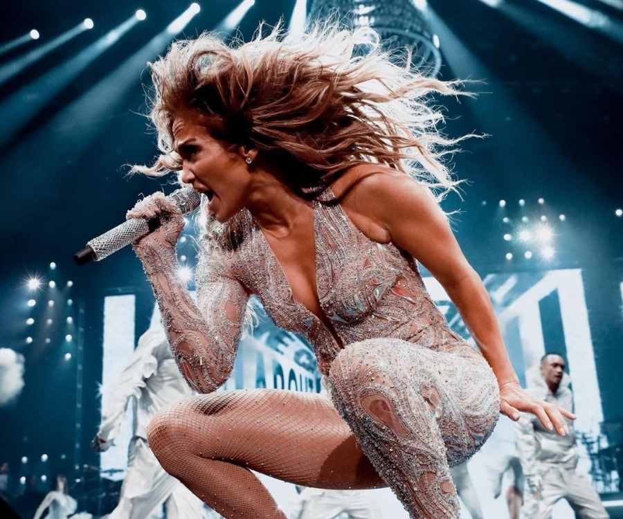 Jennifer Lopez cantando agachada em show. No fundo da foto, tem dançarinos e luzes.