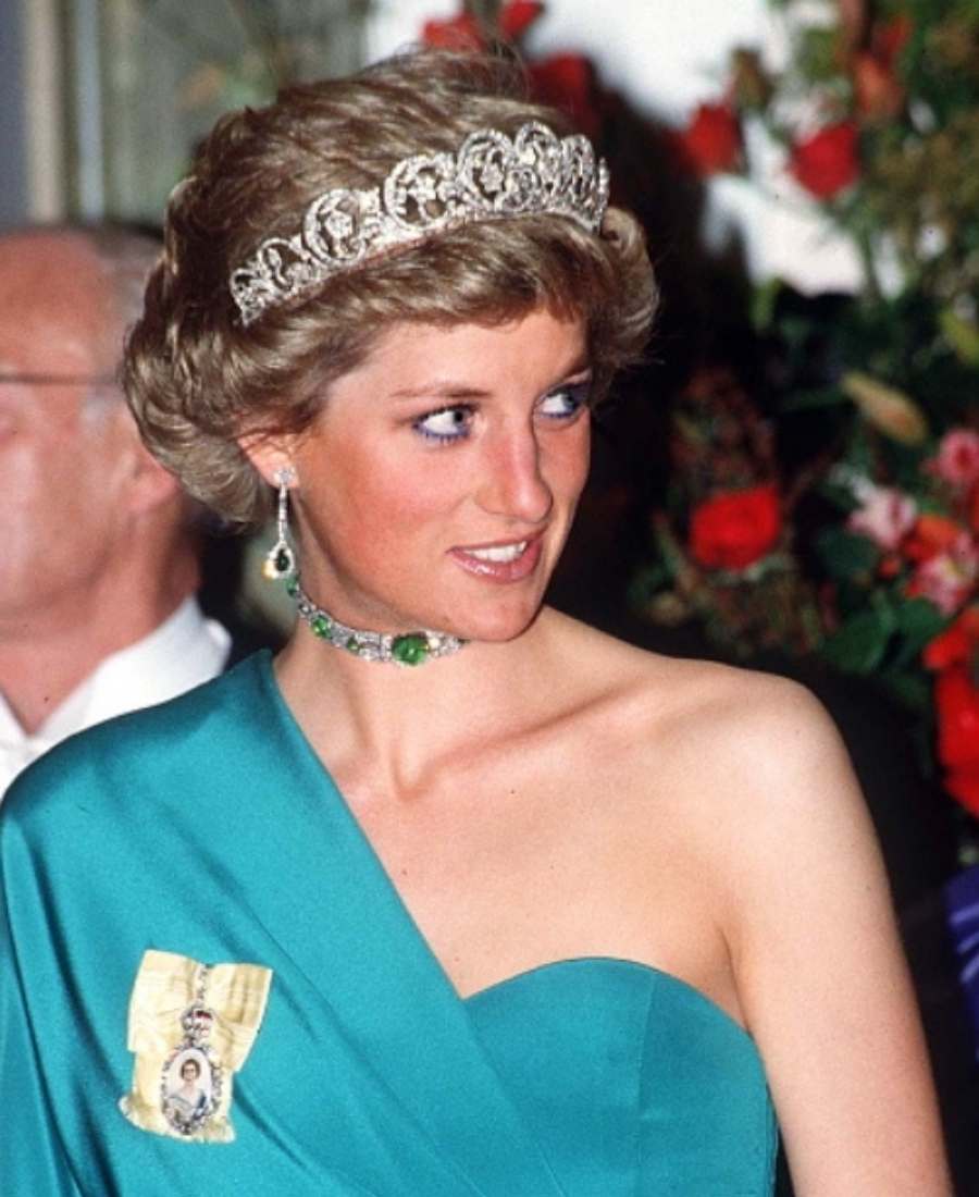 Foto da  da princesa de Galesa em um local com pessoas e decoração de flores. Ela está de frente e usa Tiara Spencer, gargantilha e brincos de esmeraldas, roupa verde-água e broche com imagem de santo. 