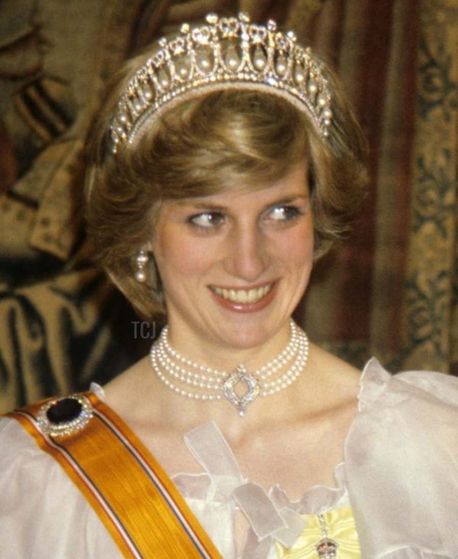 Foto da princesa Diana sorridente possivelmente no palácio de Buckingham, no Reino Unido. Ela está de frente, usando gargantilha de pérolas, Lover's Knot Tiara, brincos, roupa branca e uma faixa amarela nos ombros com broche de joia.