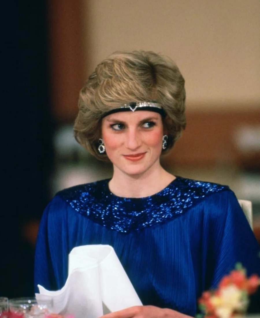 Foto  da princesa de Gales em um restaurante com mesa, copos, flores e guardanapos. Ela está de frente e usa gargantilha de diamantes na cabeça, brincos e roupa azul escuro.