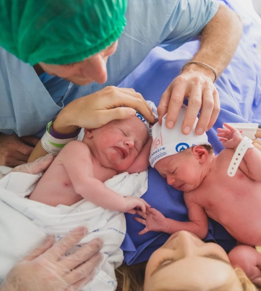 Bebês Mel e Bento no colo de Isa na maternidade. Papai Ricardo e a mamãe Isa estão com uma das mãos na cabeça dos gêmeos.