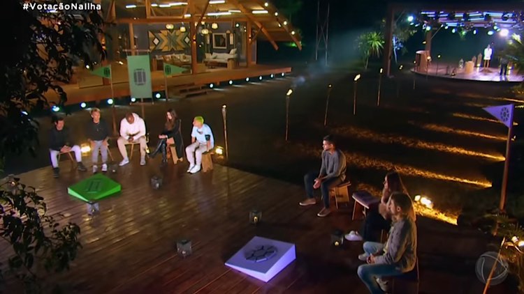 Imagem da parte externa da Vila do Ilha Record, todos os jogadores estão sentados em uma plataforma de costas para outro palco onde Mariana Rios comanda a votação.