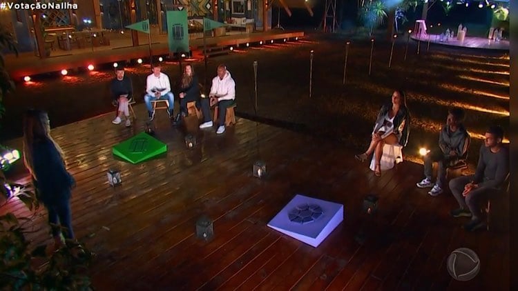 Jogadores do Ilha Record na noite de votação na parte externa da casa. Eles estão divididos por equipes, sentados em lados opostos e Mariana Rios comandando a votação.