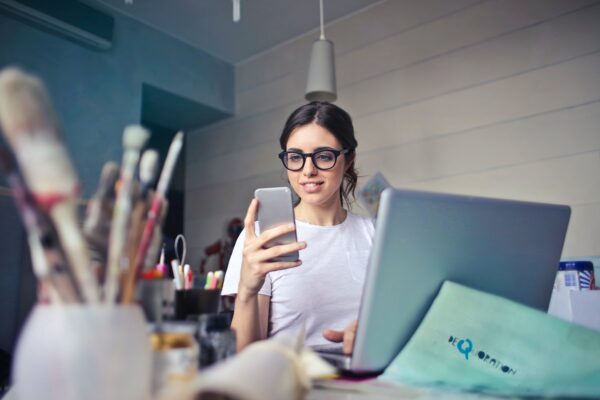 mulher de óculos sorrindo sentada em frente a um notebook e olhando para o celular