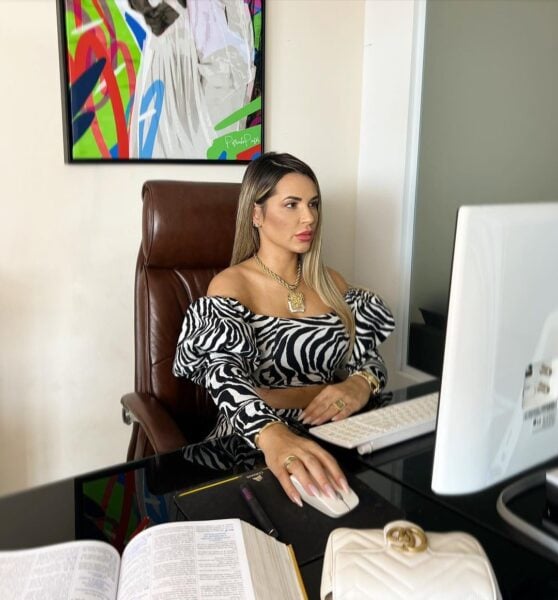 Foto da Deolane com expressão séria, sentada em frente ao computador com um livro ao seu lado, trabalhando
