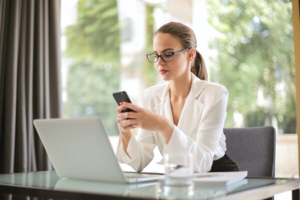 mulher de óculos com aspecto sério, sentada em frente ao notebook e usando o celular