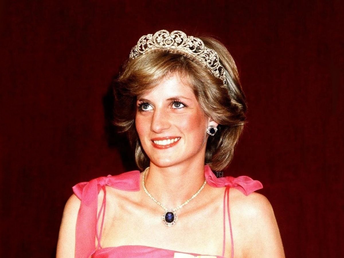 Foto de Lady Di usando joias: tiara, brincos e colar. Ela está sorridente e usando roupa pink, Atrás dela, o fundo é marrom.