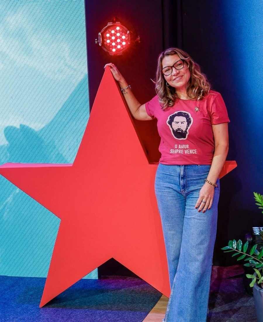 Foto da mulher do candidato à presidência sorridente e em pé ao lado de uma estrela vermelha de quase a sua altura. Ela está apoiando uma das mãos na estrela e usando camiseta com estampa do Lula e calça jeans.