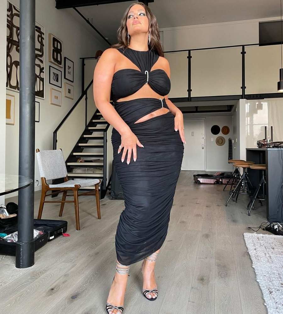 Foto de Ashley Graham. Ela está com as mãos no quadril, usando vestido preto longo de recortes. Está em casa com escada e decoração simples.