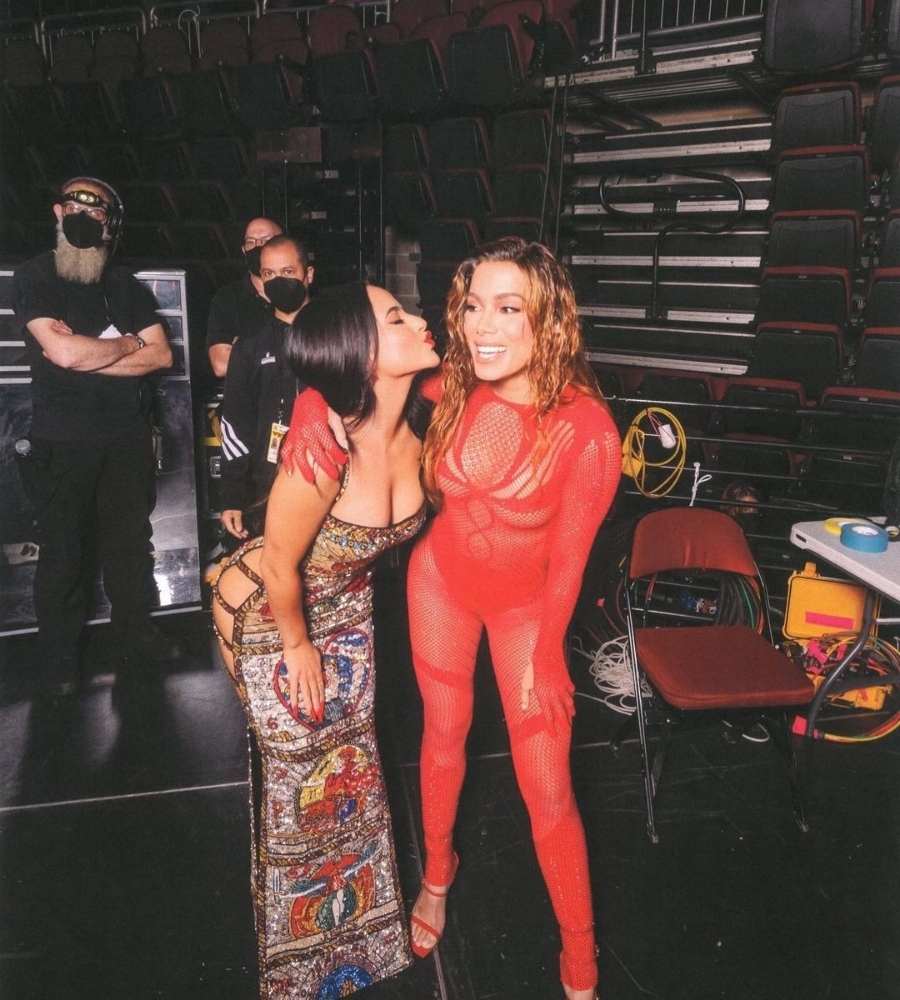 Foto de Becky G com Anitta no VMA 2022. Ela está usando vestido com recortes e pedras coloridas e beijando próximo do rosto de Anitta, que veste roupa vermelha. Ambas estão em local com 3 rapazes, mesa, cadeira, fios e outros objetos dos bastidores.
