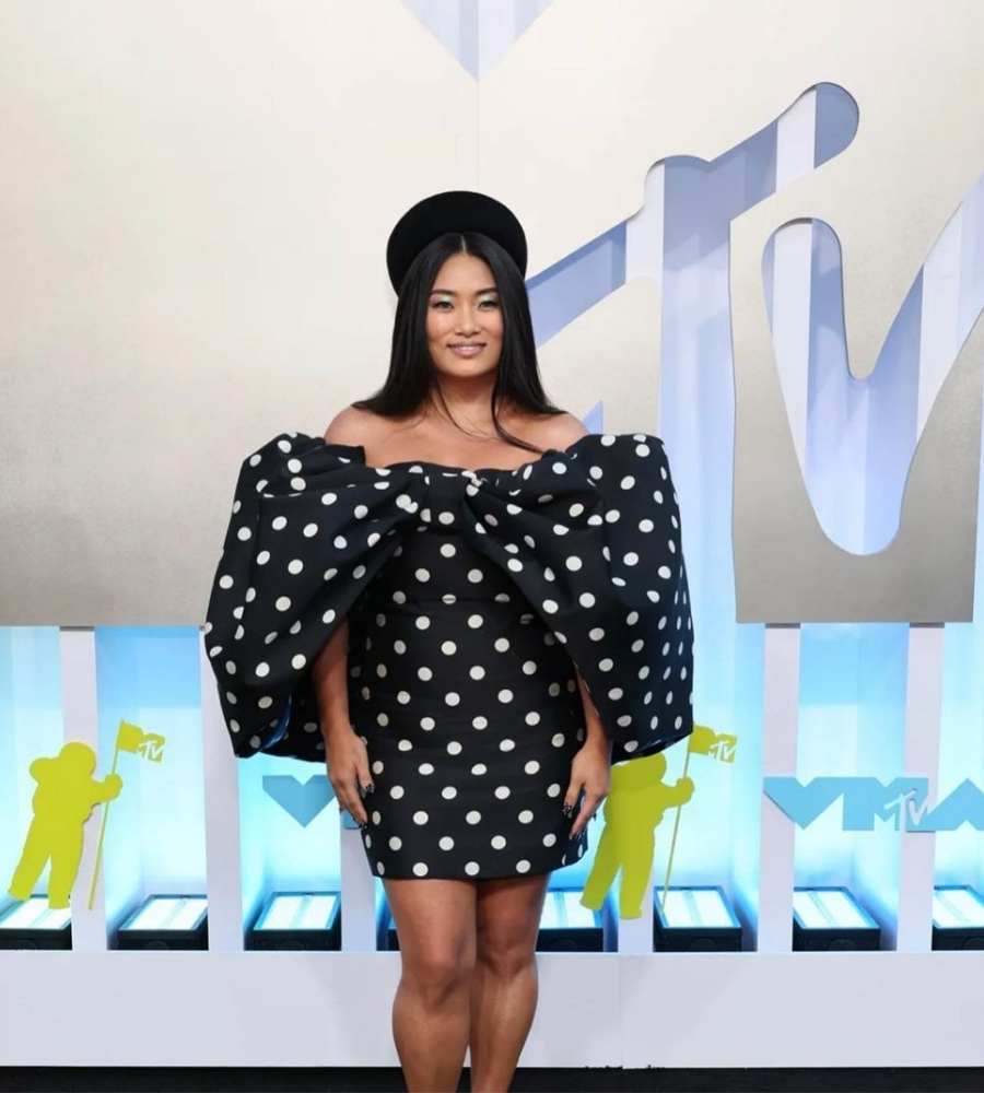 Foto de Chloe Flower no tapete do evento. Ela está sorridente, usando vestido curto preto de bolinha branca. Usa também boina preta.