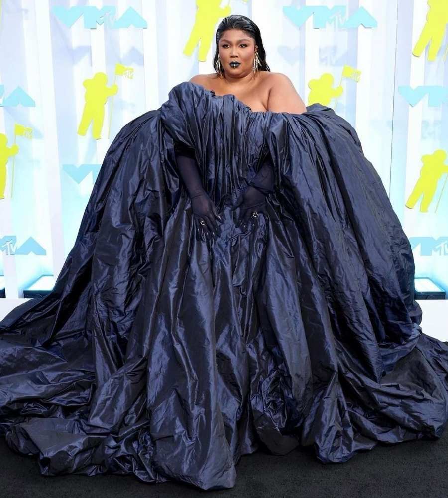 Foto de Lizzo no tapete evento. Ela usa vestido preto mega volumoso e ainda luvas.