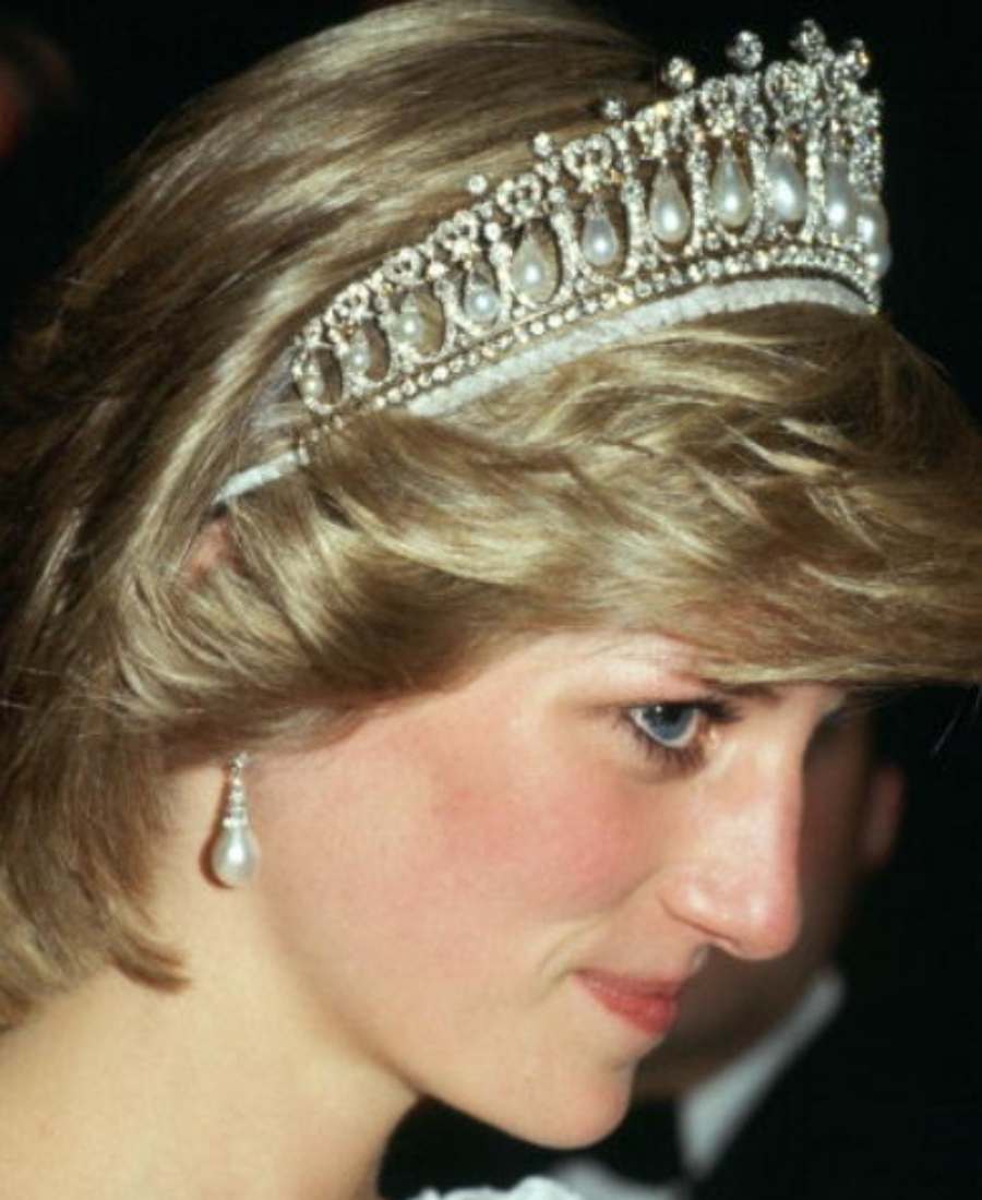 Foto do rosto da princesa Diana em um fundo preto. Ela está de perfil e usa Lover's Knot Tiara e brincos. Atrás dela, é possível ver a silhueta de um homem.