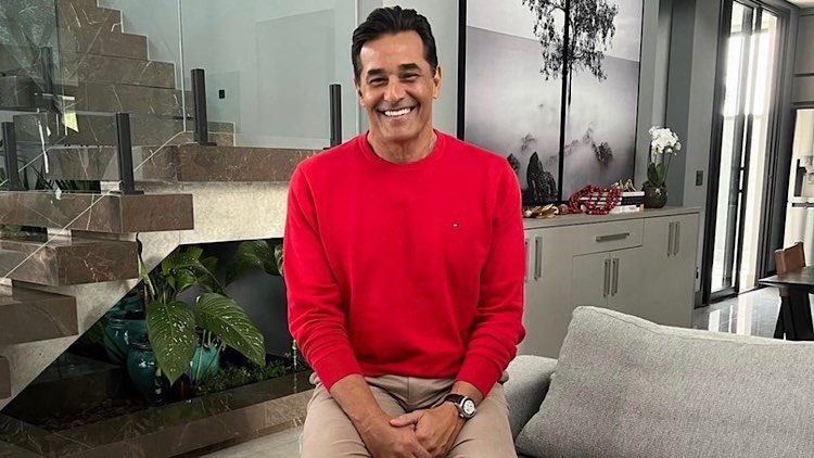 Luciano Szafir sentado em sua sala sorrindo de blusa vermelha.
