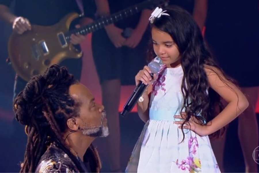 Foto da atriz mirim e Carlinhos Brown. Ela está com microfone e ele agachado olhando para ela. Os dois estão no palco do programa.